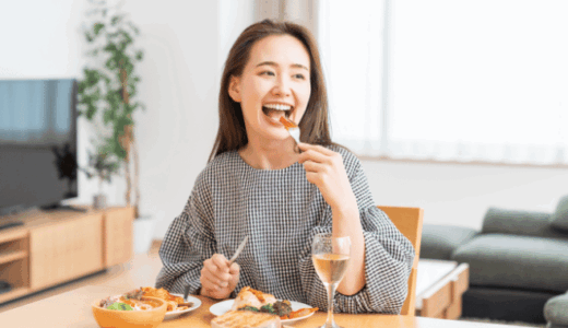 宅配弁当を楽しそうに食べる健康志向の女性。木製のテーブルに栄養バランスの取れた食事が並び、笑顔でフォークを口に運んでいる様子