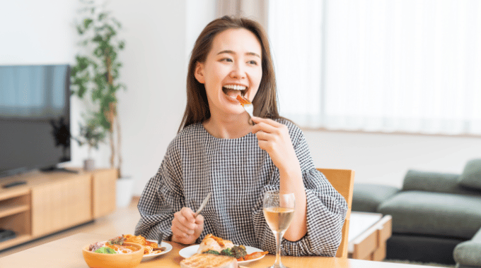 宅配弁当を楽しそうに食べる健康志向の女性。木製のテーブルに栄養バランスの取れた食事が並び、笑顔でフォークを口に運んでいる様子